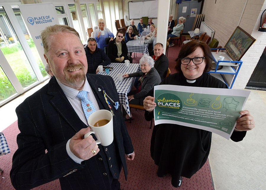 Councillor Paul Dean and Reverend Lesley Jones with people who are sat at tables with drinks.